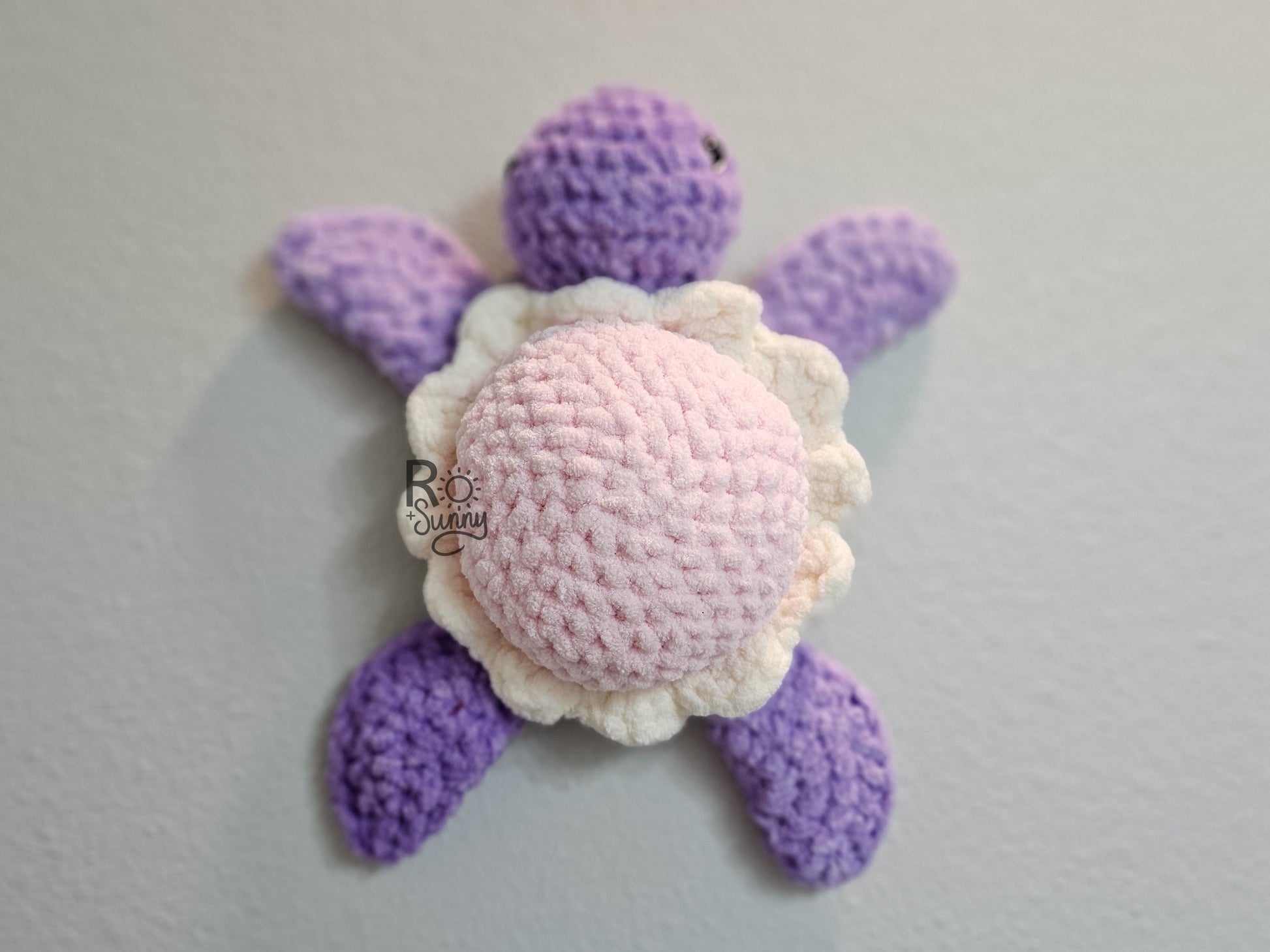 Cotton Candy, Overhead, Lying Flat  - Crochet sea turtle with a purple body and light pink shell with that has a white ruffle. The belly of the turtle is a light teal color. 