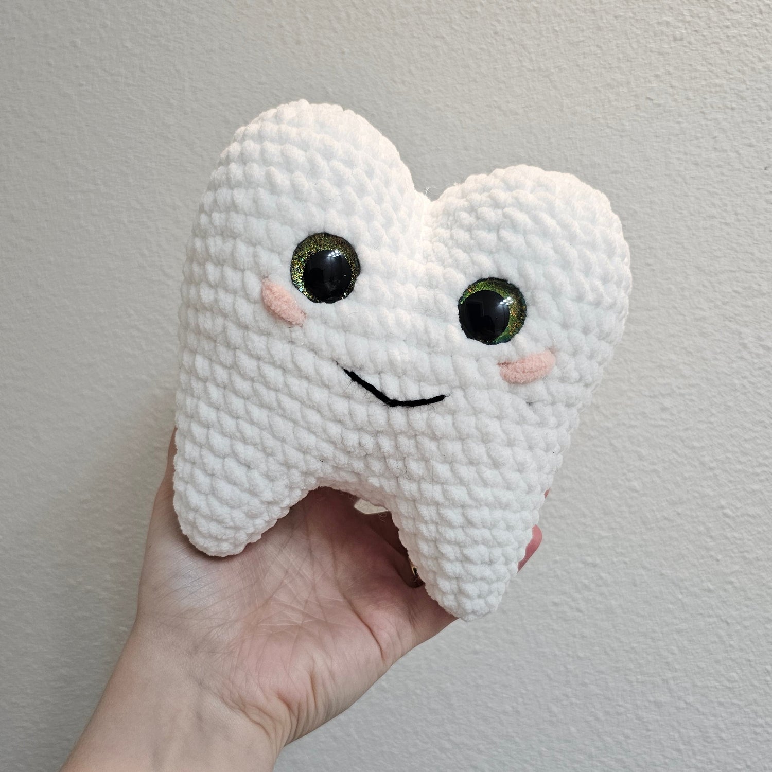 Hand holding a white crochet tooth with black eyes and a smile against a plain background