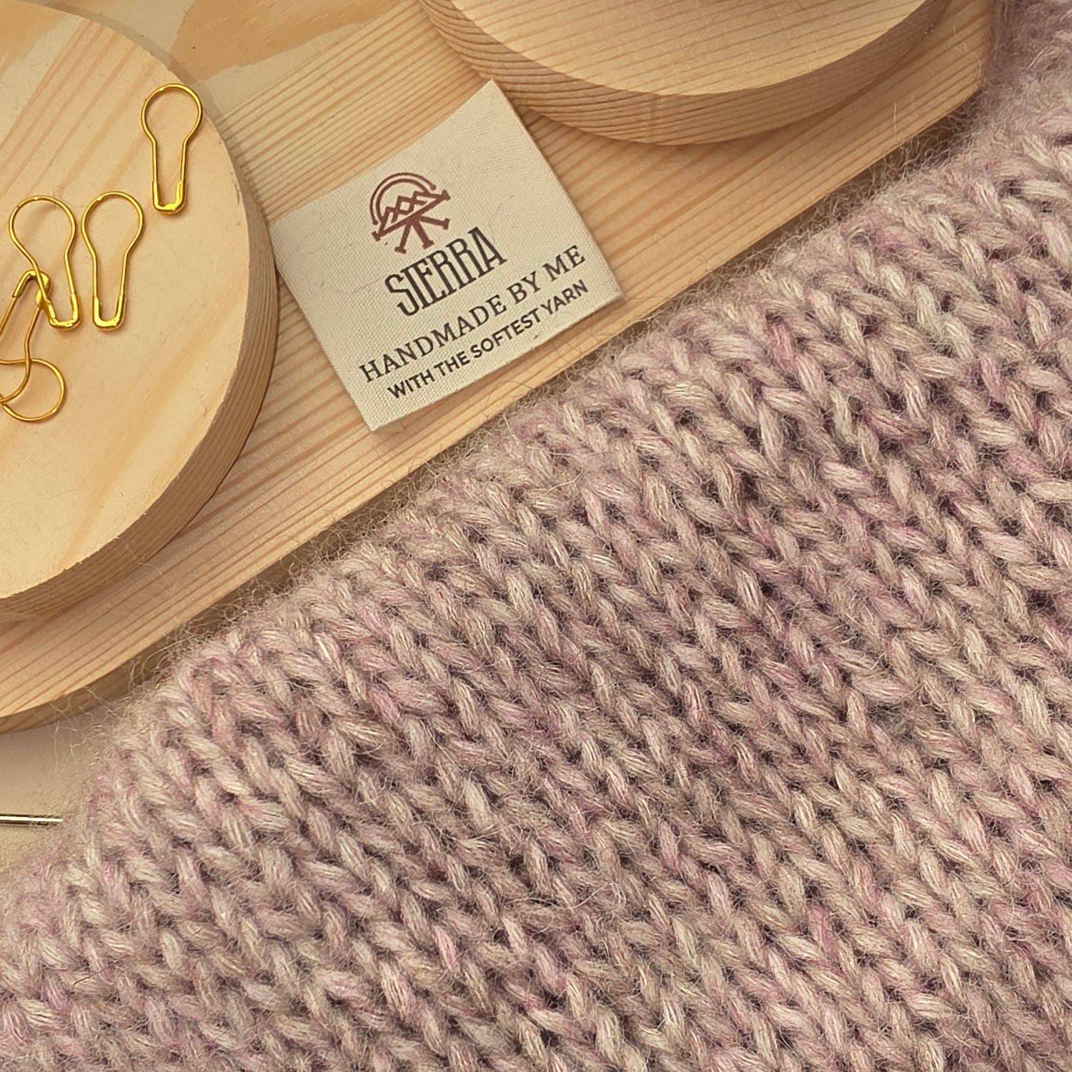 Close-up of a knitted fabric in a soft dusty pink color, with a small fabric tag reading 'SIERRA YARN HANDMADE BY ME' and wooden display stands visible in the background.